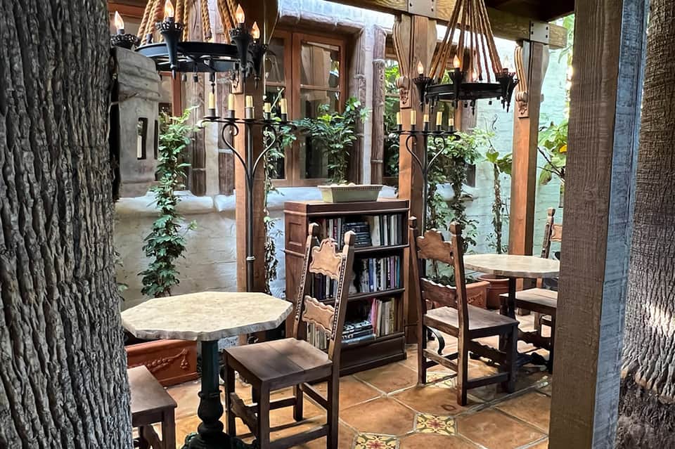 Spanish colonial courtyard with stone columns, wooden tables, hanging chandeliers, and lush greenery