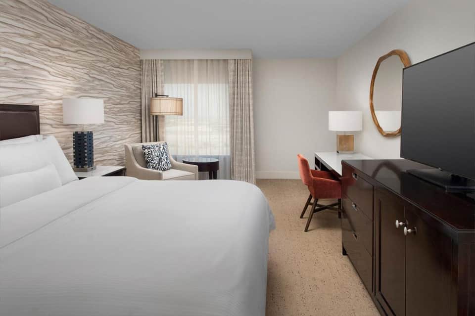 Spacious hotel bedroom with king bed, modern furnishings, textured accent wall, and desk area