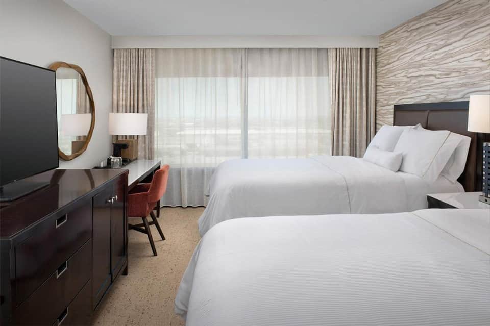 Modern hotel room with two queen beds, dark wood furniture, and floor-to-ceiling curtains