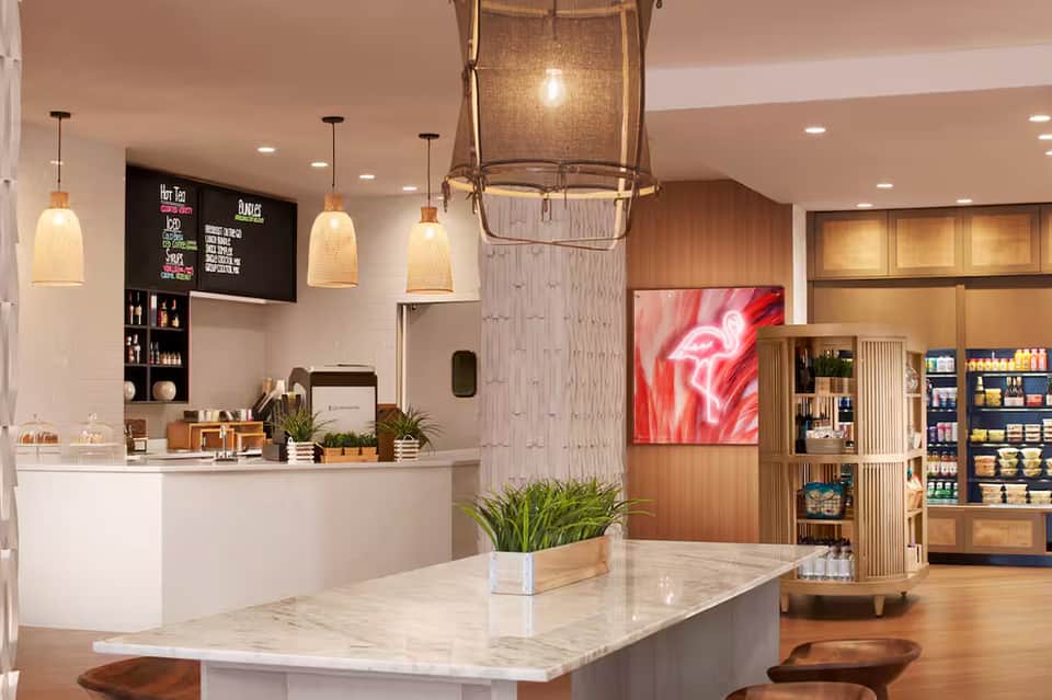 Modern hotel lobby with white marble counter, pendant lights, wooden shelving, neon signage, and indoor plants