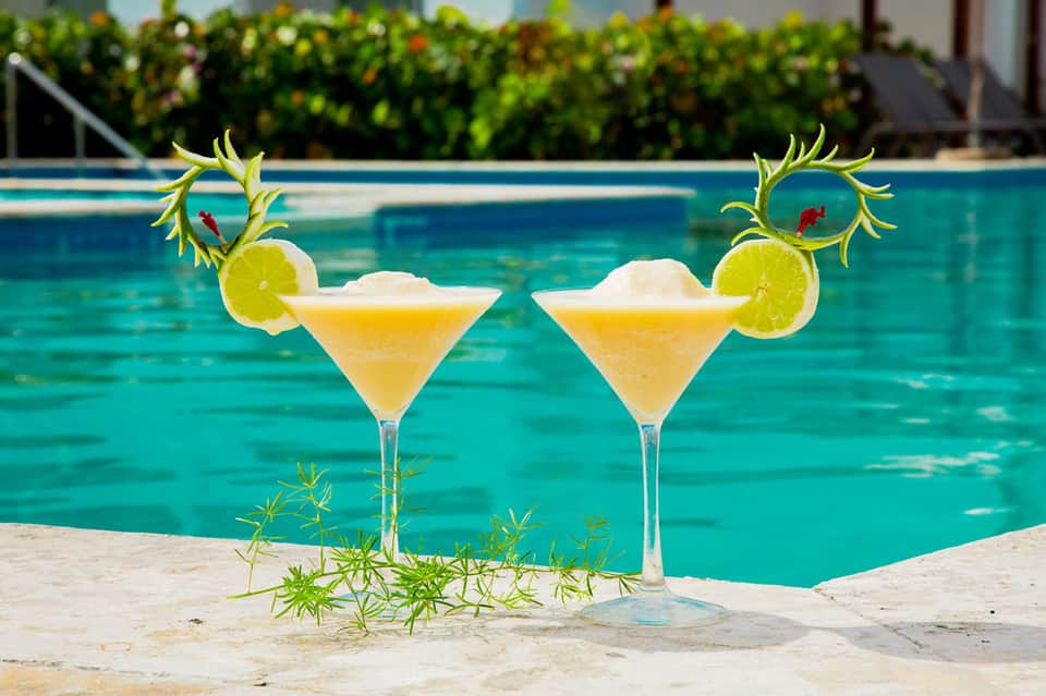 Two frozen margaritas with lime garnish positioned poolside with turquoise water in background