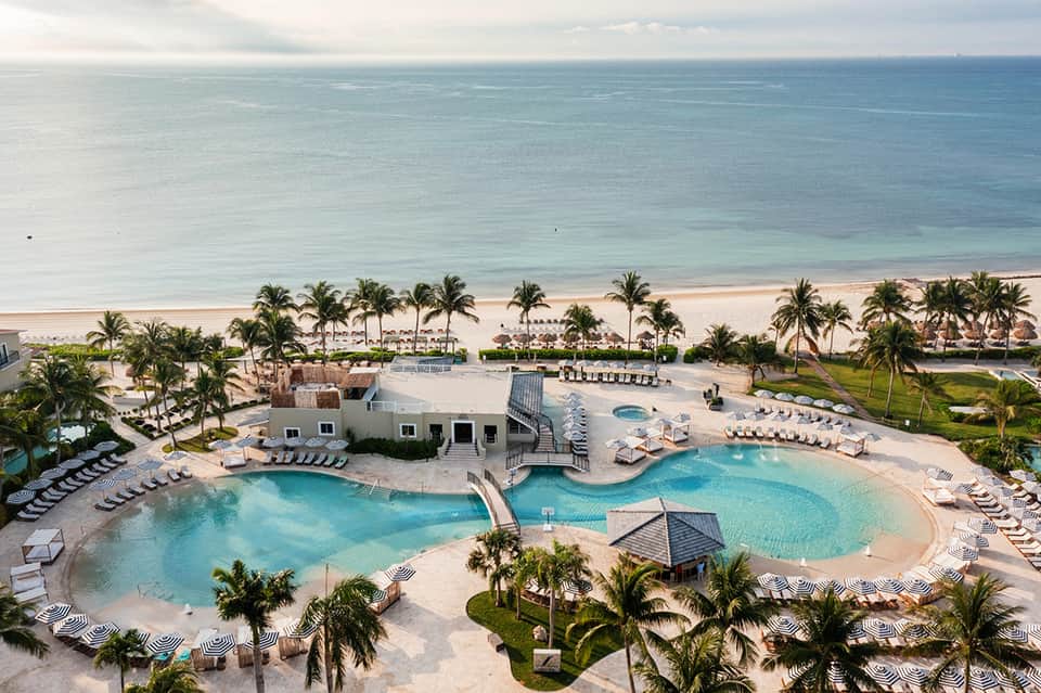 Aerial view of beachfront resort with turquoise pool, palm trees, loungers, and ocean beyond