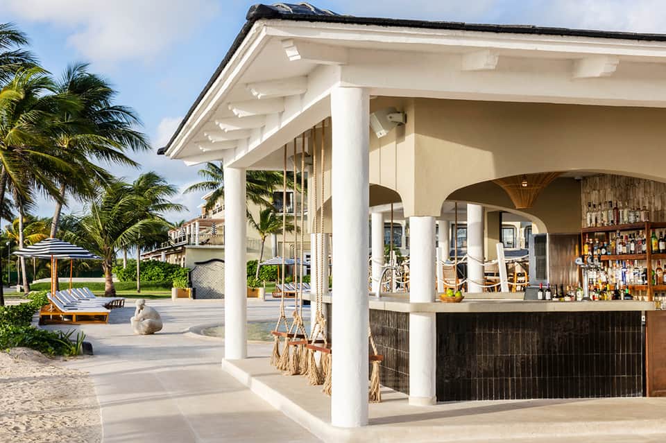 Beachfront bar pavilion with white columns, dark wood counter, and palm trees under blue sky
