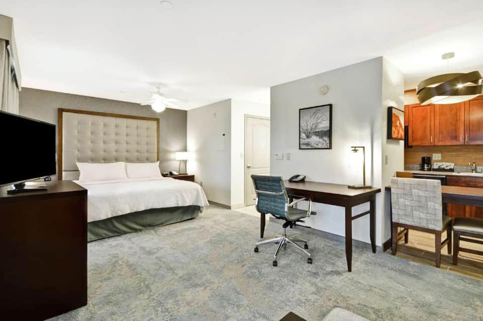 Hotel suite bedroom with queen bed, work desk, kitchenette, and contemporary furnishings