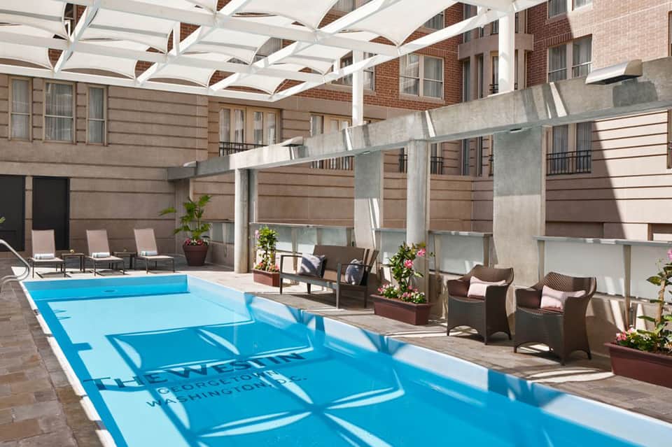 Outdoor hotel pool with blue water, white pergola overhead, lounge chairs, and brick building facade