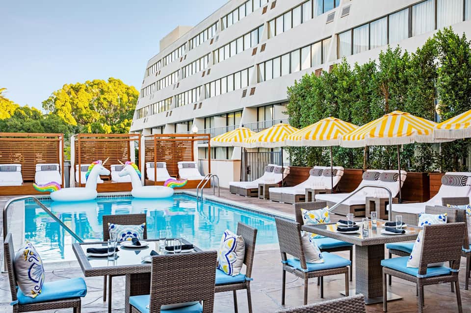 Resort pool deck with yellow-striped umbrellas, lounge chairs, dining tables, and modern white building backdrop