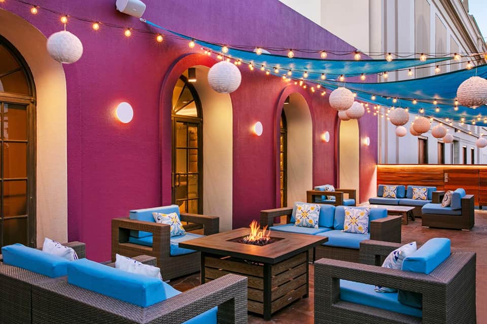 Vibrant outdoor patio with magenta walls, string lights, white lanterns, blue cushioned seating, and central fire pit