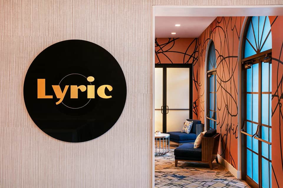 Lyric hotel entrance with orange accent walls, black circular logo sign, and modern lounge seating visible through glass doors