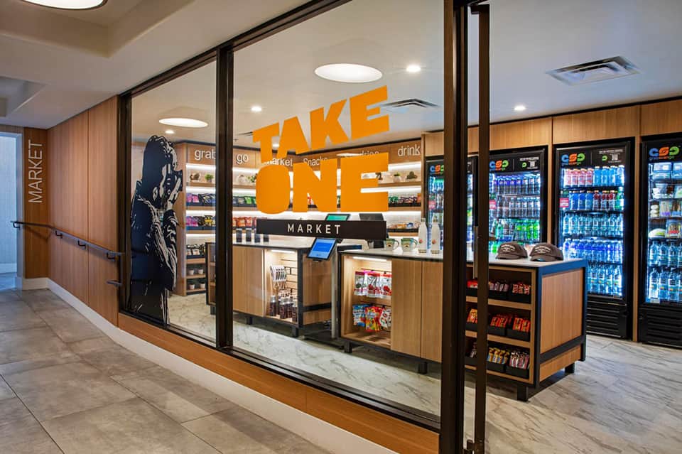 Modern hotel market convenience store with orange signage, refrigerated beverage cases, and wooden interior design