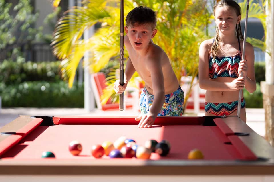 Two children playing pool at a resort, with colorful balls on the red pool table and tropical plants in background