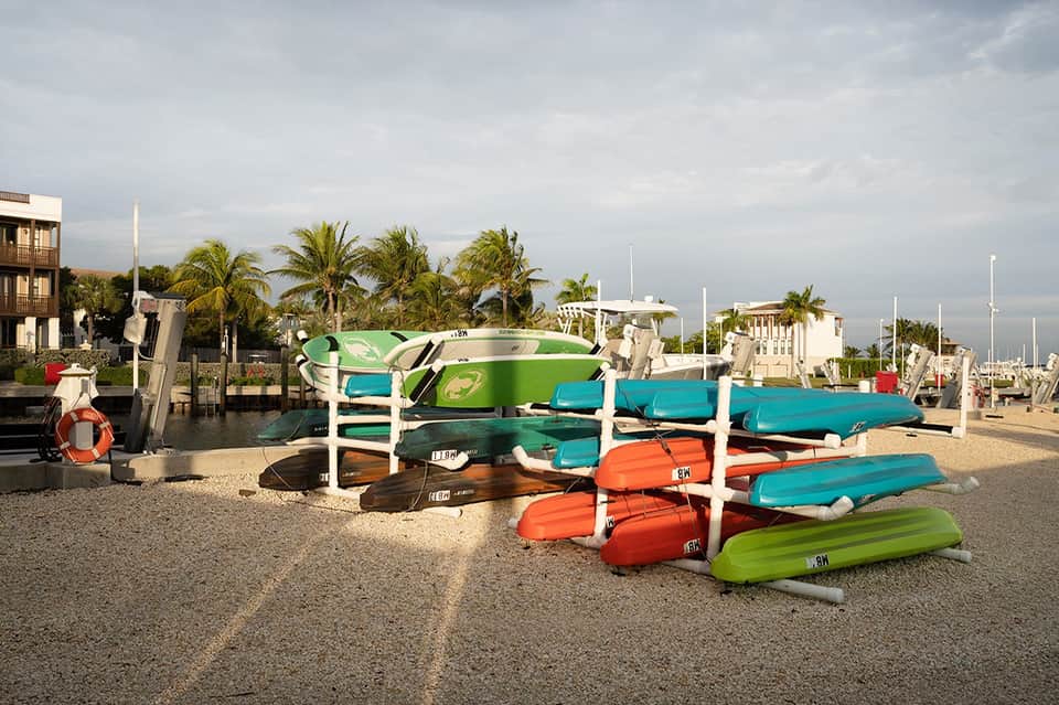 Colorful kayaks and paddleboards stacked on beach near marina with palm trees and waterfront buildings