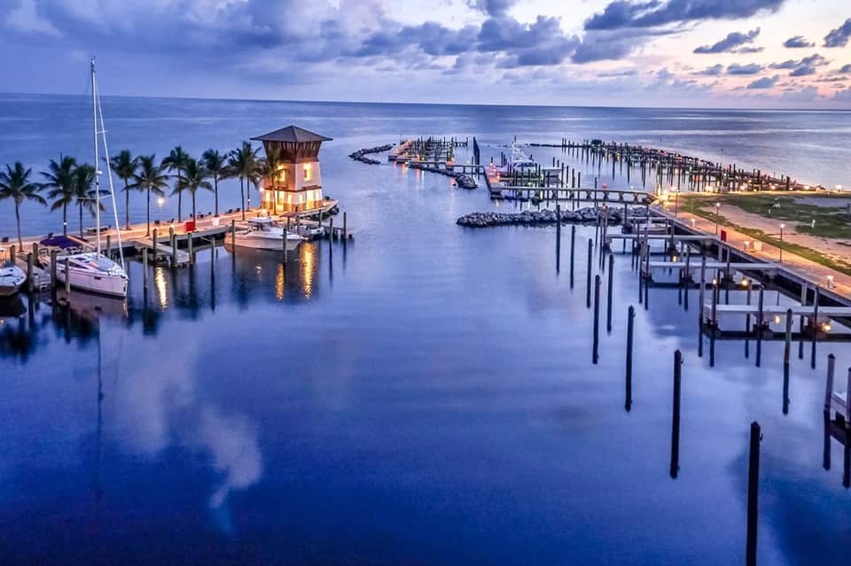 Illuminated harbor tower and wooden boat docks reflecting in calm blue water at dusk with palm trees