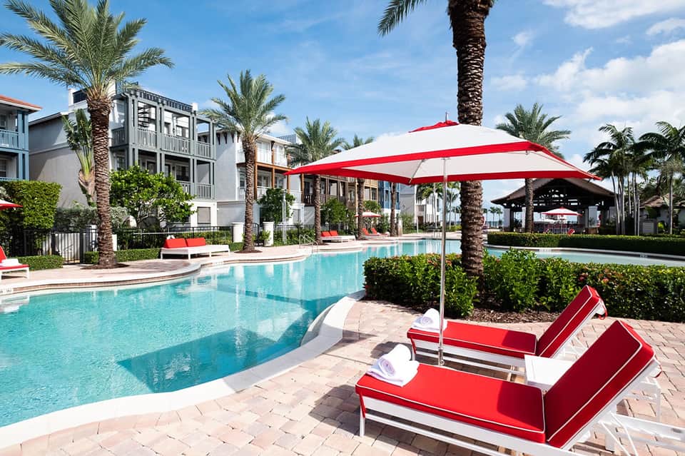 Outdoor resort pool with red and white umbrellas, loungers, and palm trees surrounding modern residential buildings
