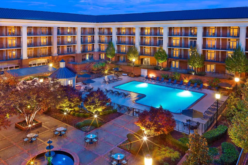 Illuminated courtyard with glowing pool, fire pit, and multi-story brick hotel building at dusk