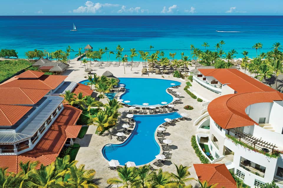 Aerial view of beachfront resort with curved blue pools, palm trees, and orange-roofed buildings overlooking turquoise ocean
