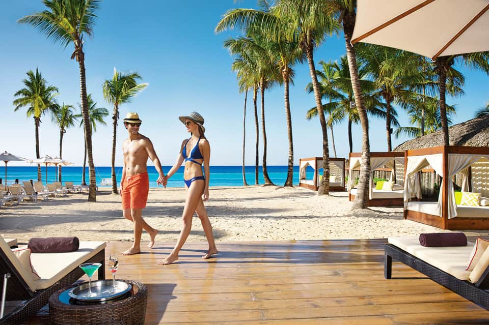 Couple walking on wooden deck with palm trees, beach lounge chairs, and ocean view at tropical resort