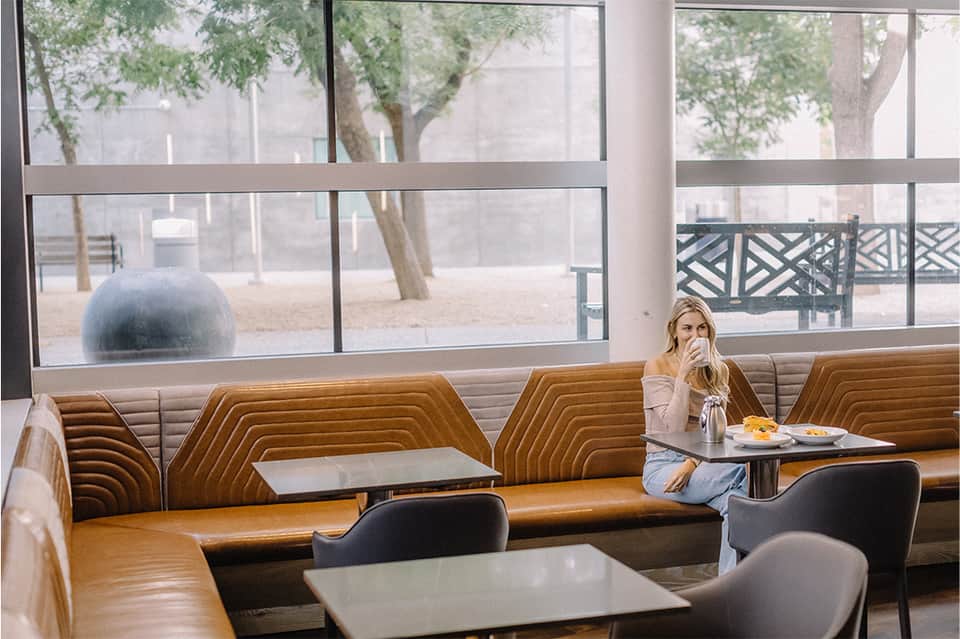 Contemporary café seating area with tan leather booth seating, small tables, and large windows overlooking an urban plaza