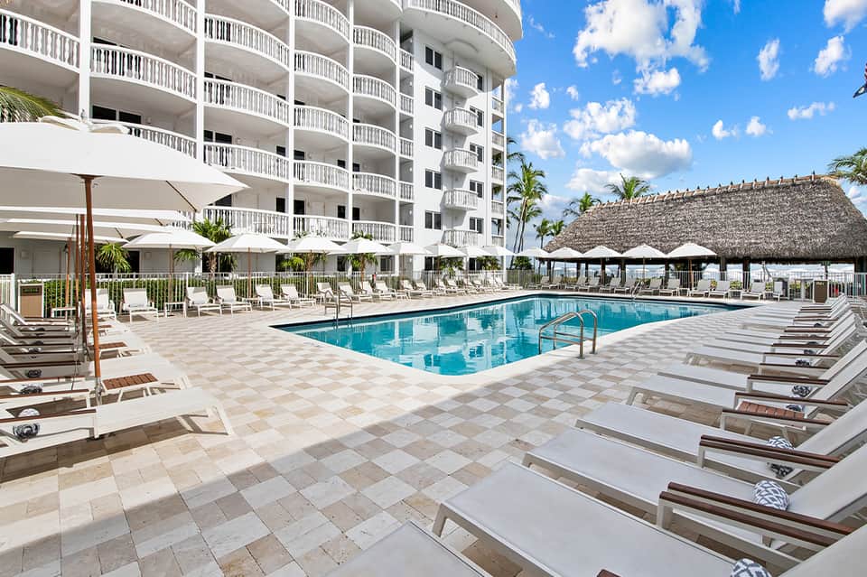 Resort pool with white loungers, umbrellas, and multi-story building with balconies and thatched roof