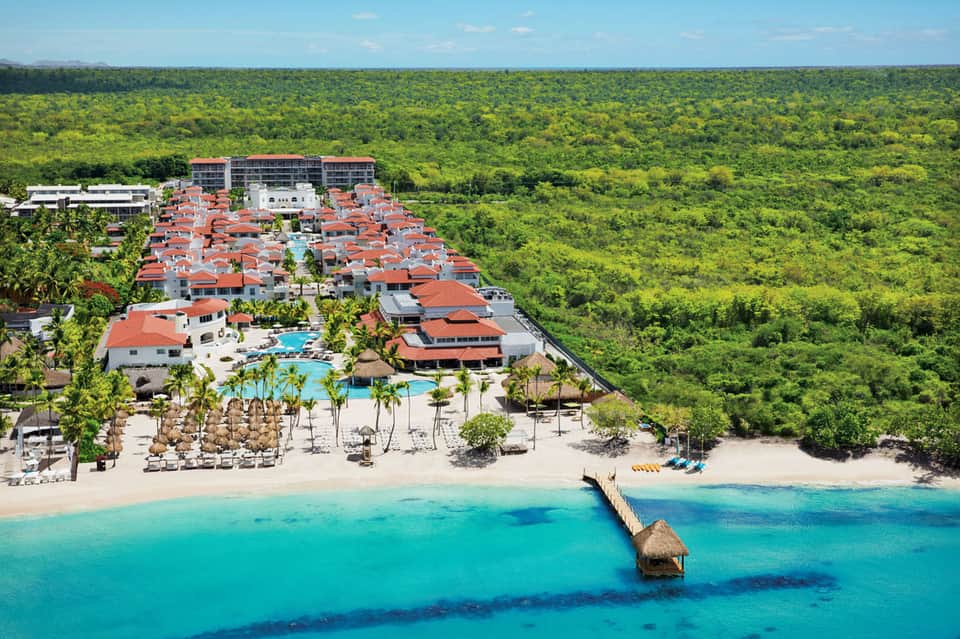 Beachfront resort with red-roofed villas, pool, and pier overlooking turquoise Caribbean waters surrounded by jungle