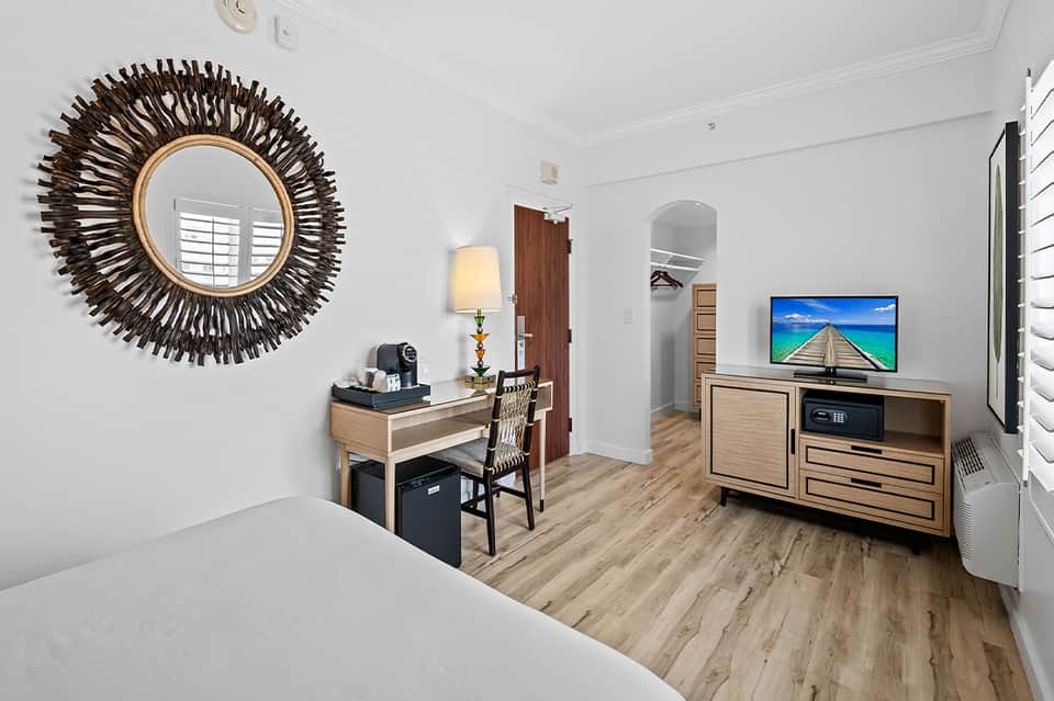 Hotel suite with queen bed, desk, TV, decorative sunburst mirror, and wood flooring