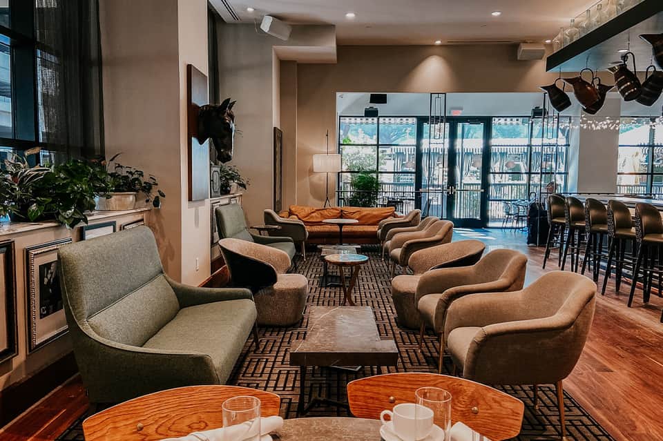Modern hotel lounge with orange sofa, tan armchairs, wooden tables, and floor-to-ceiling windows overlooking a balcony