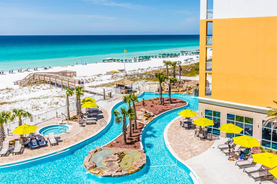 Beachfront resort with curved lazy river pool, yellow umbrellas, and pristine white sand beach with turquoise water
