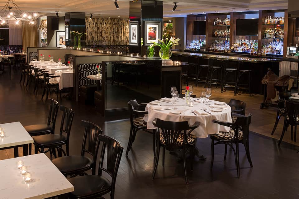 Elegant fine dining restaurant with round tables, black chairs, white linens, and full bar with bottles