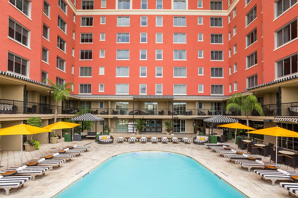 Coral-red courtyard building with striped lounge chairs, blue pool, yellow umbrellas, and black-and-white accent details