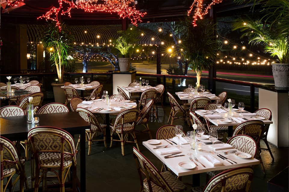 Outdoor restaurant terrace at night with rattan chairs, white tablecloths, string lights, and city views