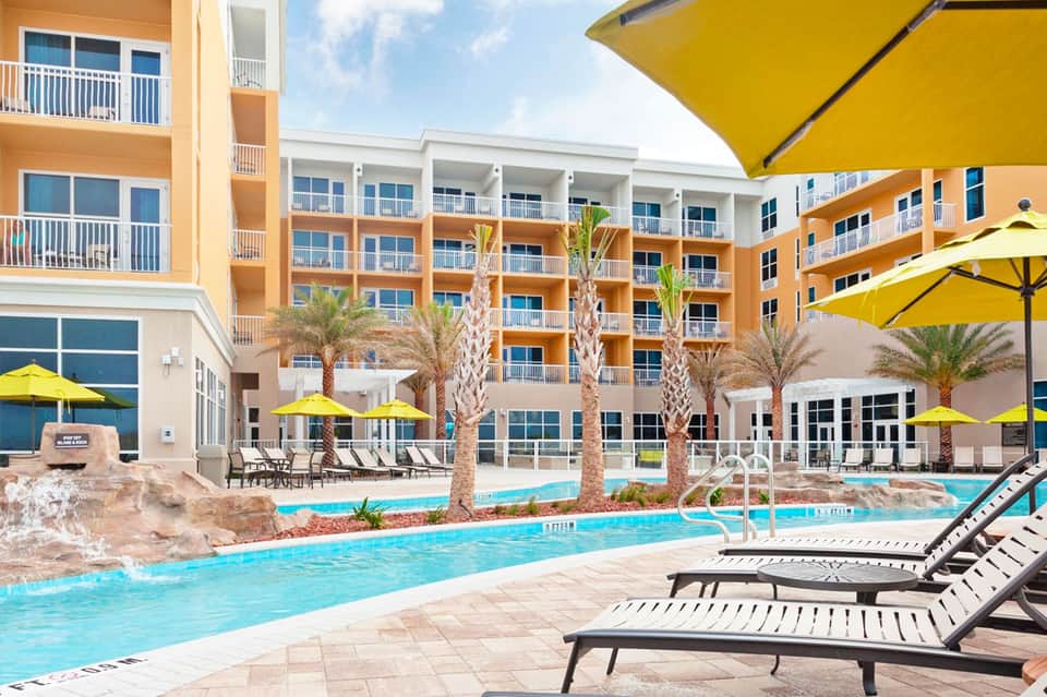 Resort courtyard with orange buildings, palm trees, yellow umbrellas, and curved swimming pool with lounge chairs