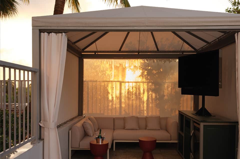 Covered outdoor lounge area with beige sofa, cushions, side tables, and mountain view at sunset