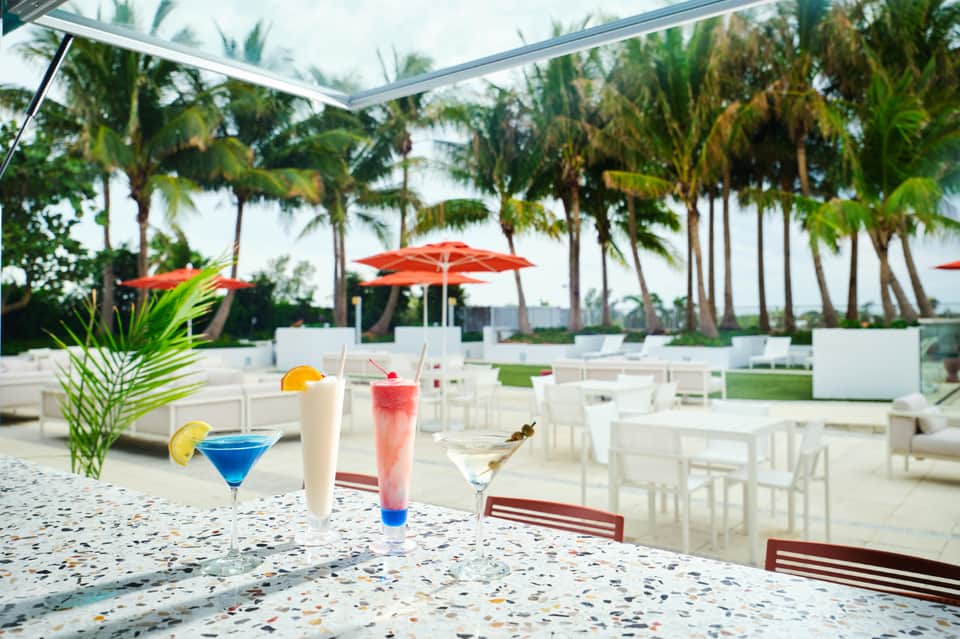 Tropical resort bar and lounge with colorful cocktails, palm trees, and red umbrellas overlooking ocean