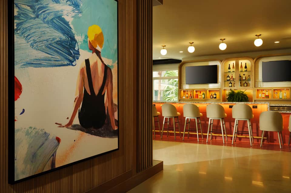 Modern hotel bar lounge with orange seating, illuminated shelving, and contemporary artwork featuring a beach scene