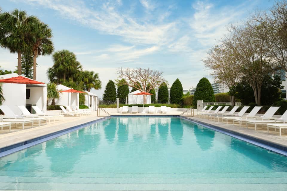 Resort pool with clear turquoise water, white lounge chairs, red umbrellas, and palm trees under blue sky
