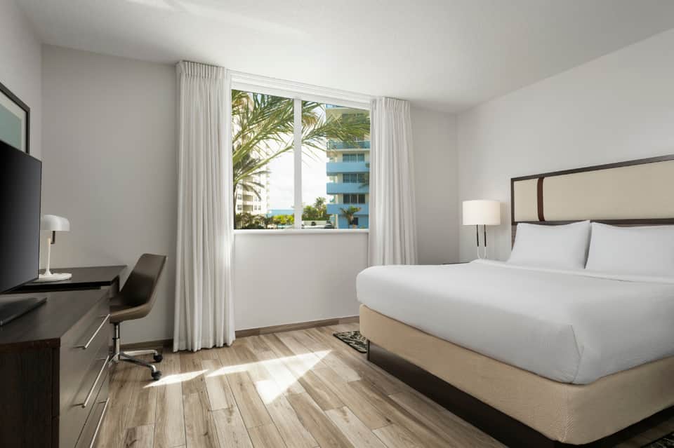 Modern hotel bedroom with ocean view through large windows, white bedding, dark wood desk, and light wood flooring