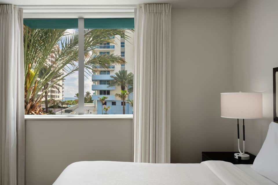 Beachfront hotel room with ocean view through large windows, palm trees visible, white bedding, and minimalist décor