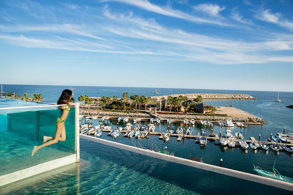 Infinity pool overlooking marina with boats, palm trees, and rocky breakwater