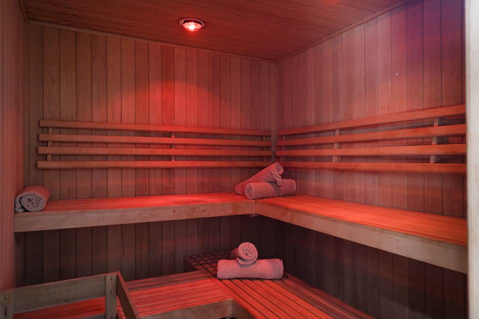 Infrared sauna interior with wooden benches, rolled towels, and red heat lamp illuminating the cedar-lined room