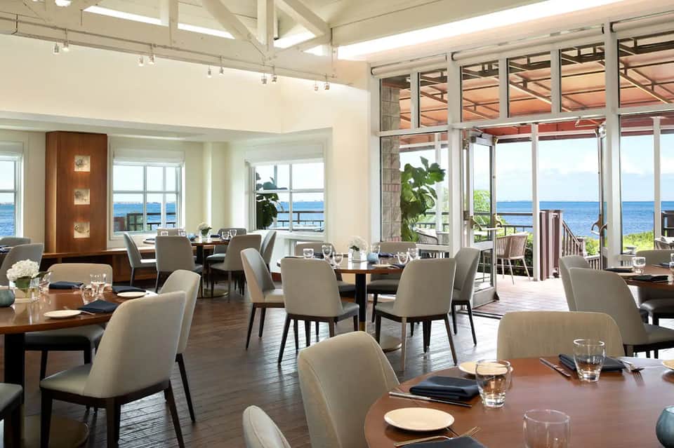 Elegant coastal dining room with floor-to-ceiling windows overlooking the ocean, wooden tables, beige chairs, and modern lighting