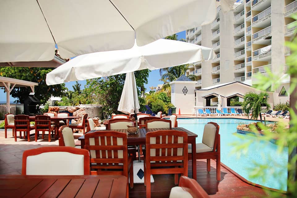 Poolside dining area with red wooden chairs, white umbrellas, and multi-story resort building in background