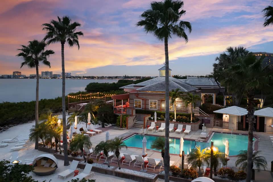 Luxury waterfront resort at dusk with illuminated pools, palm trees, and buildings overlooking calm bay waters