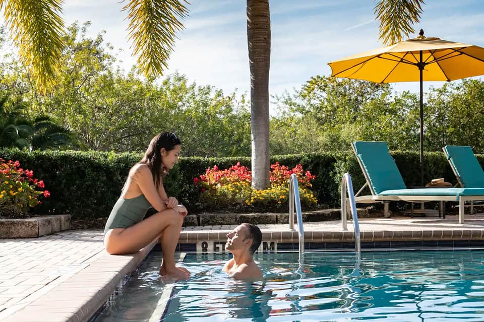 Woman in pool talking to person on deck beside yellow umbrella and turquoise lounge chairs