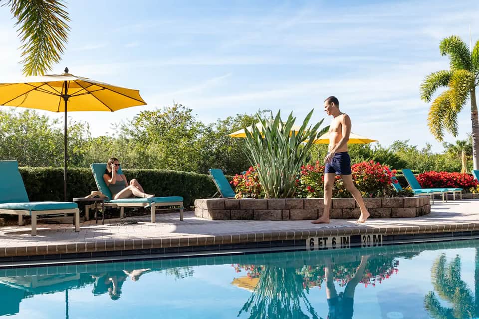 Resort pool deck with turquoise loungers under yellow umbrellas, palm trees, colorful flowers, and person about to dive