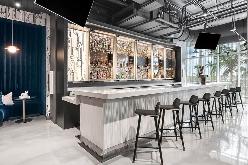 Modern hotel bar with illuminated glass shelving, white bar counter, black bar stools, and industrial ceiling