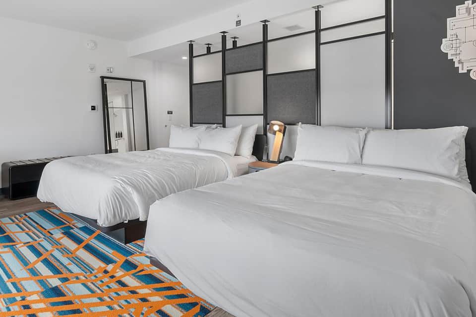 Contemporary hotel room with two queen beds, dark headboard, geometric patterned floor rug, and minimalist design