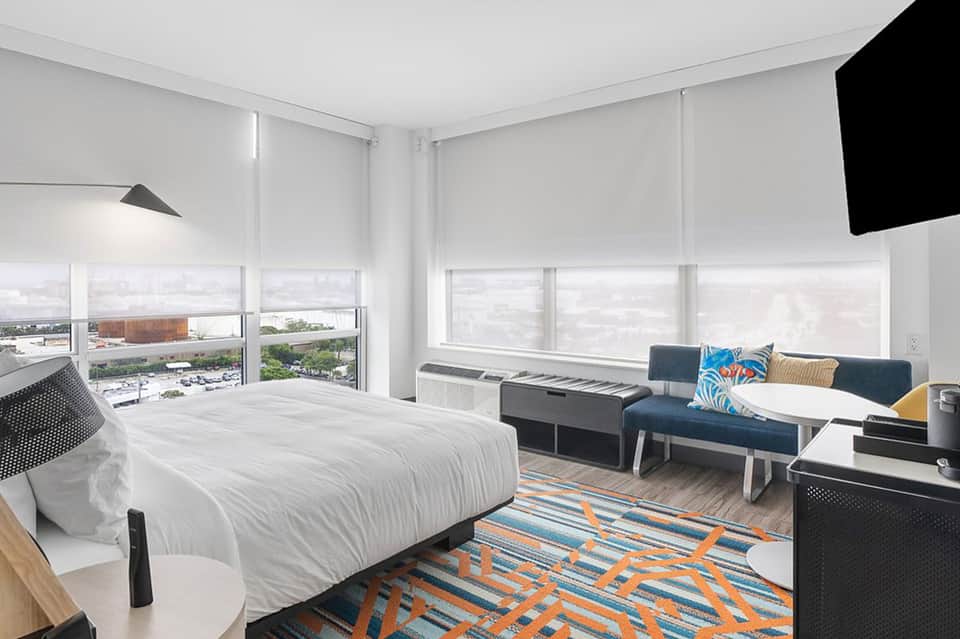 Modern hotel room with white bedding, blue sofa, expansive city views, and geometric area rug
