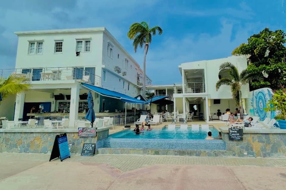 Beachfront resort with white buildings, pool area, palm trees, and blue umbrellas
