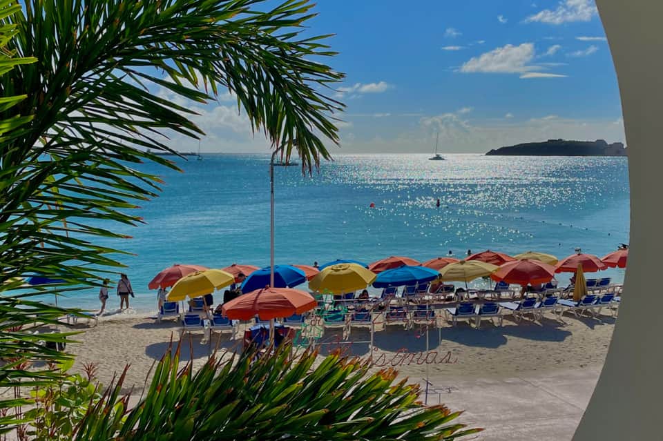 Colorful beach umbrellas line sandy beach with turquoise ocean and sailboats under clear blue sky