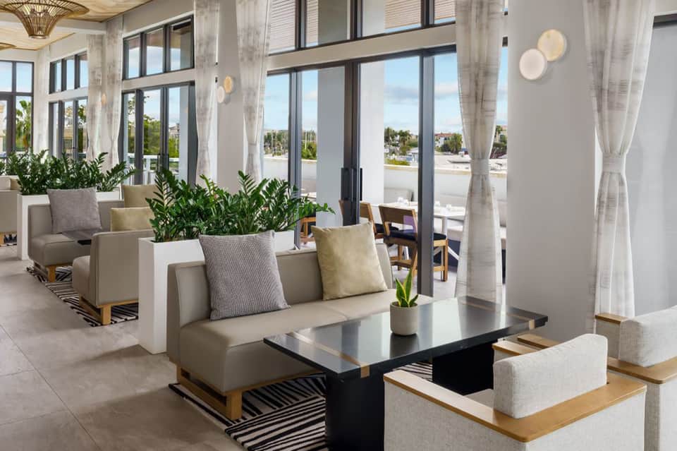 Bright, modern lounge with beige sofas, large windows overlooking city, indoor plants, and contemporary light fixtures