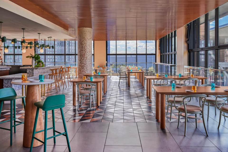 Modern high-rise restaurant with floor-to-ceiling windows, wooden ceiling, turquoise bar stools, and panoramic city views